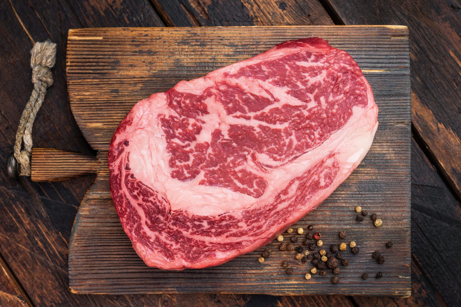 Raw, heavily marbled ribeye steak on a wooden board with colourful peppercorns